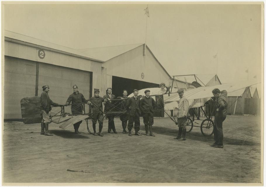 Group of 9 men stand in front of a monoplane. Background: hangars made of corrugated metal. Woman sits in the cockpit with a bulldog.