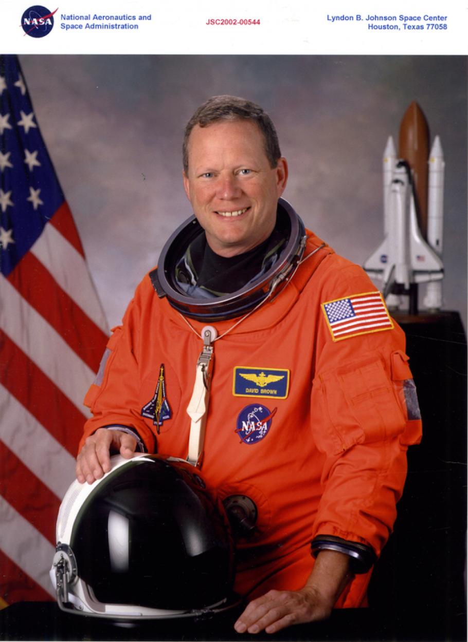Portrait photograph of man with short sandy brown hair, wearing an orange astronaut jumpsuit with patches (left in shape of shuttle, upper right nameplate "David Brown," bottom right NASA globe logo, American flag on left sleeve.  Right hand resting on helmet.  Blurred background: American flag on left, shuttle model on right.