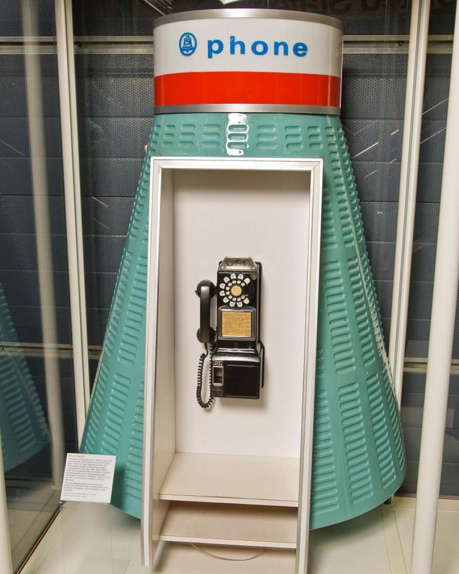 A phonebooth in turquoise color that is shaped like a space capsule with a dial phone in the middle.