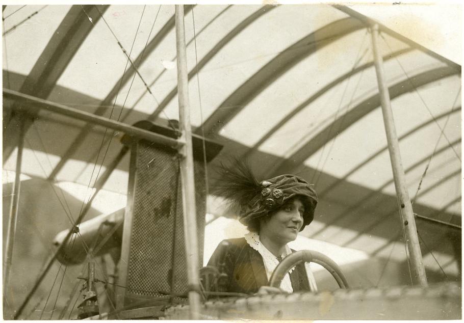 A woman in a hat with three pins and a feather behind a steering wheel.