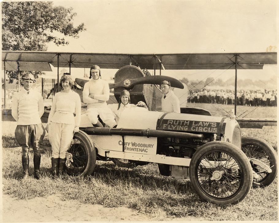 Five people lounge on a car labeled "Ruth Law's Flying Circus." Behind the car is an airplane.