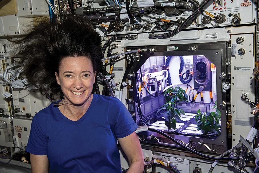 Astronaut smiling in front of plants in lit cubby on space craft.