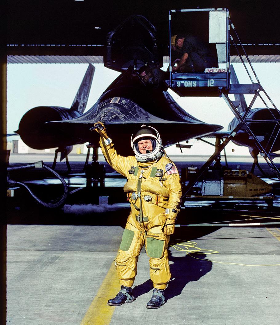 A man in a yellow suit and a space helmet points to the Blackbird airplane behind him. 