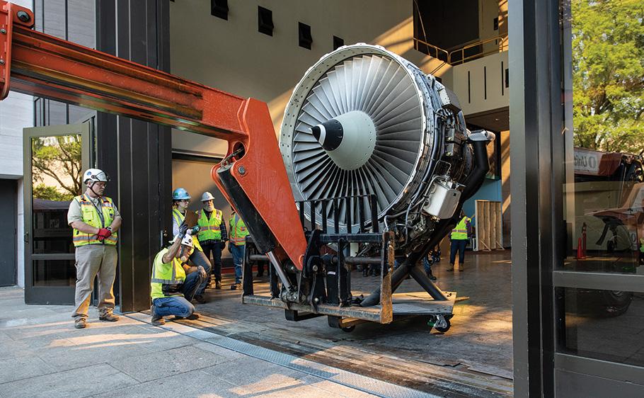 Eight workers in hard hats supervise the turbofan being moved by a large mechanical arm