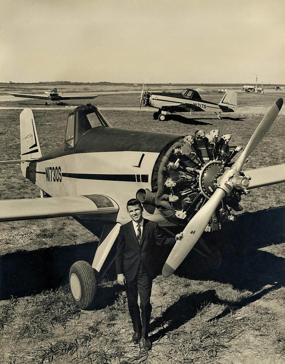 A man stand next to a small plane, his hand resting on the propeller. Two similar planes are seen in the background.