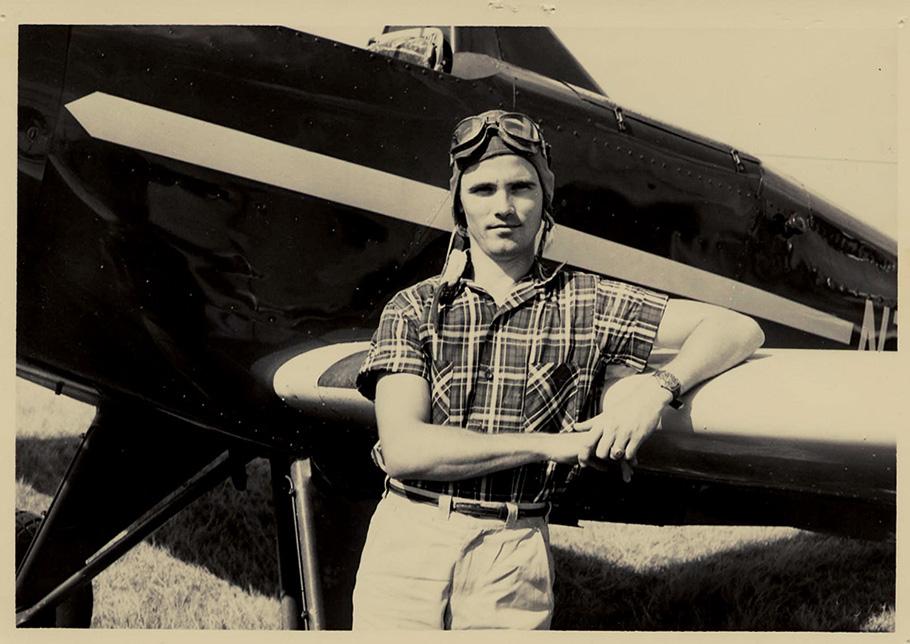 A sepia tone photograph of a man in a pilot's cap leaning against the wing of an airplane. 