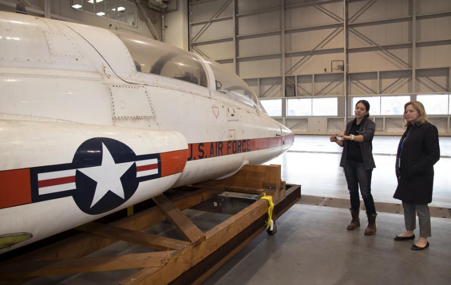 Two women examine a T-38 Talon aircraft while it's strapped to the back of a truck without its landing gears.
