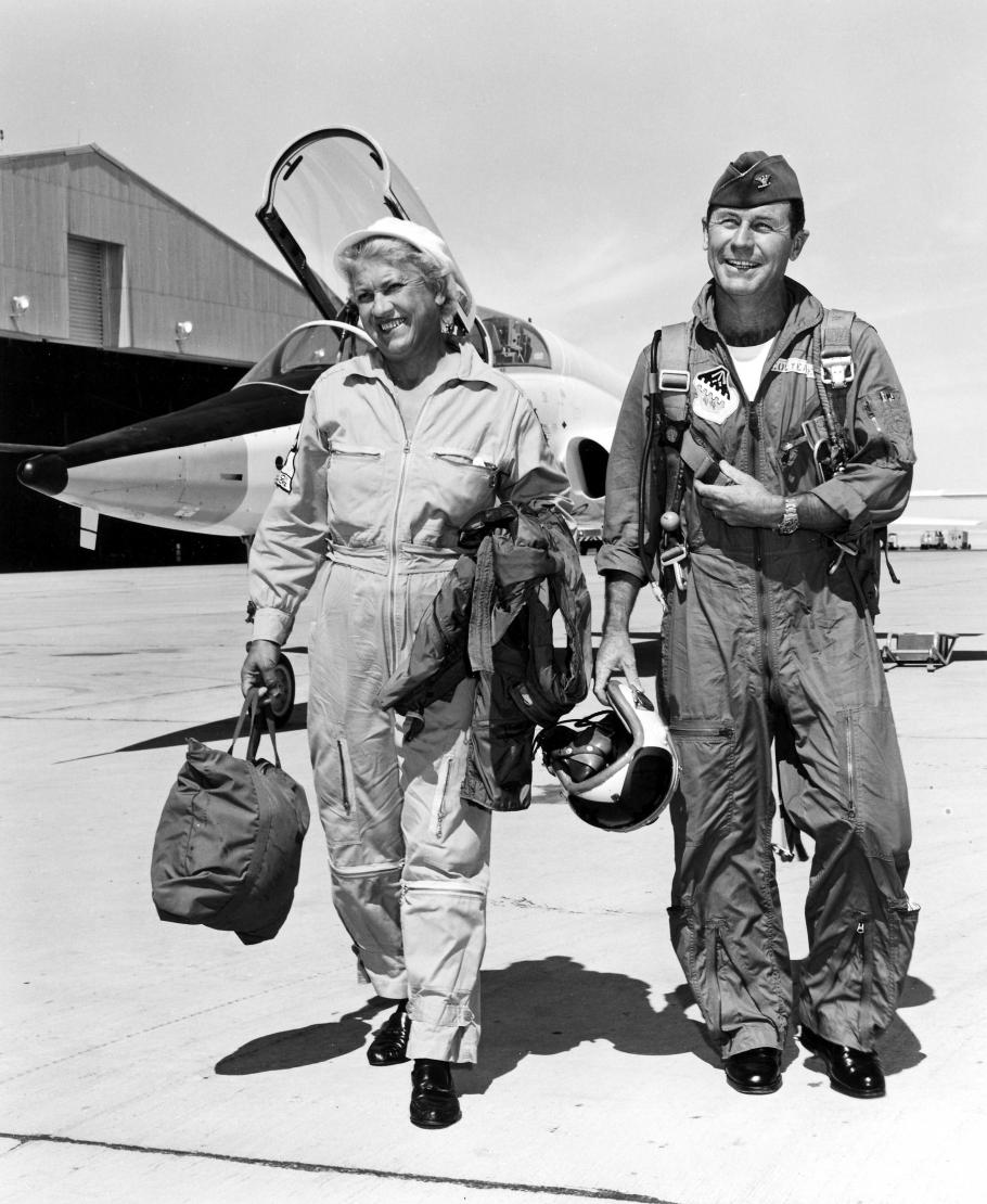 Black and white image of Jackie Cochran and Chuck Yeager walking and smiling with an aircraft in the background.