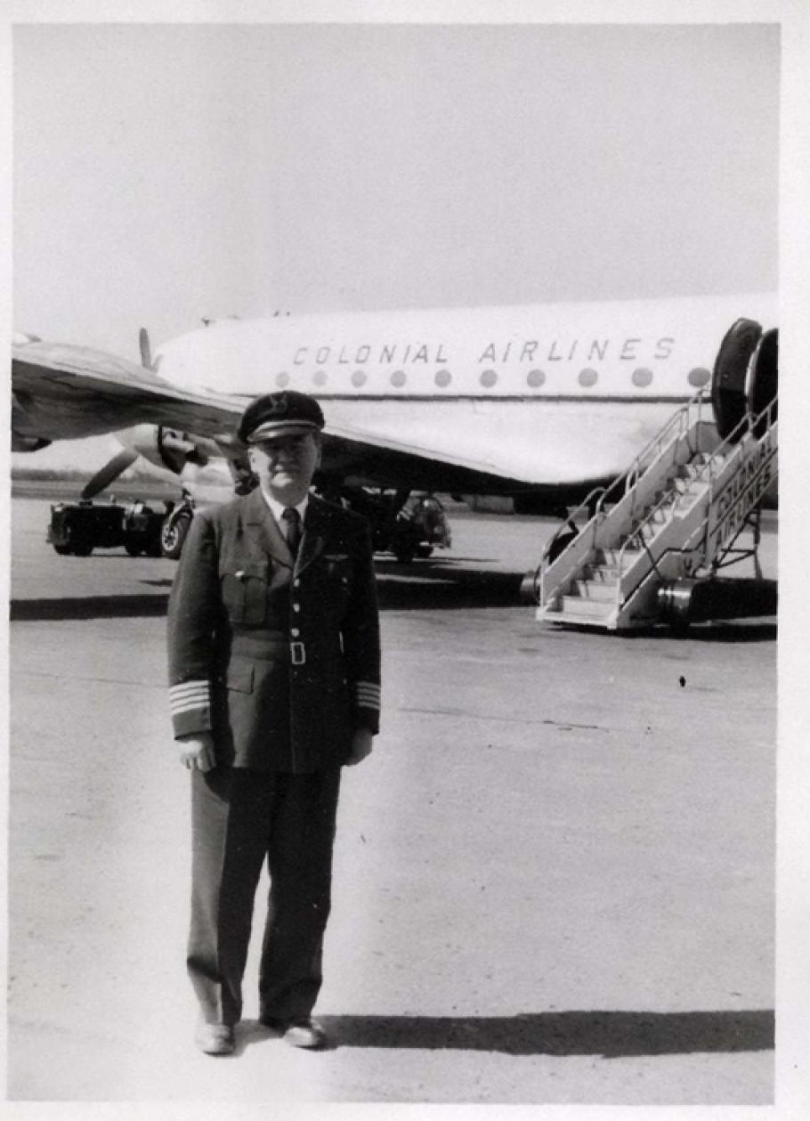 Black and white photograph. Foreground: Man in dark pilot's uniform with a cap and stripes at the ends of his sleaves. Background: Side of a passenger airplane labelled "Colonial Airlines" above circular windows. A set of airstairs is behind and to the right of the man.