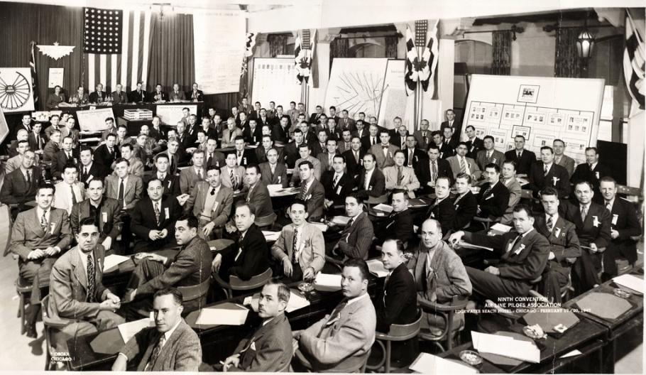 Black and white photograph of a convention of men in a large room, seated at tables. In the background on left is a podium with an eagle symbol on it and an American flag hanging vertically with the stars on the left.