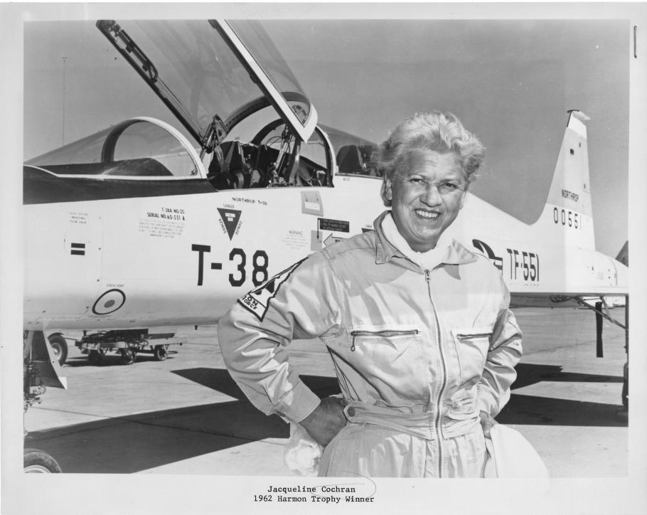 An aged Jackie Cochran posing with her hands on her hips and smiling and a T-38 Talon aircraft in the background.