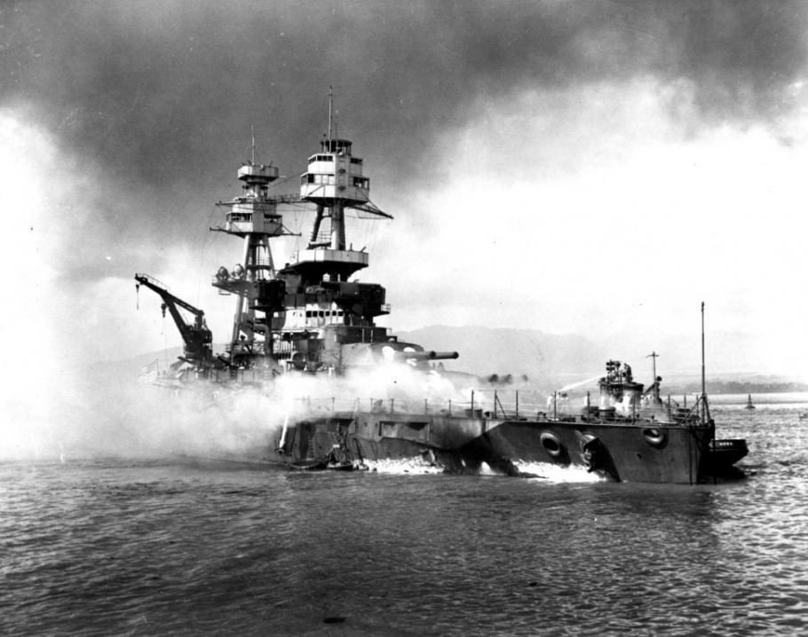 Black and white image of a battleship in flames and smoke as sits on a body of water.