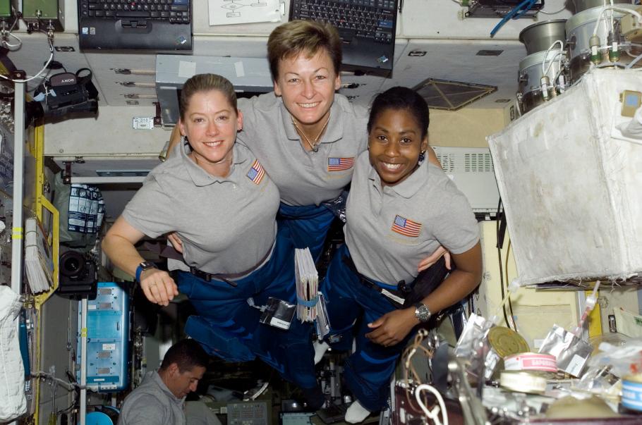 Three women locking arms and smiling while floating in zero gravity in a space station.