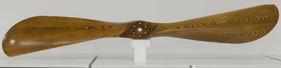 A varnished wood propeller on a white stand against an off-white background.