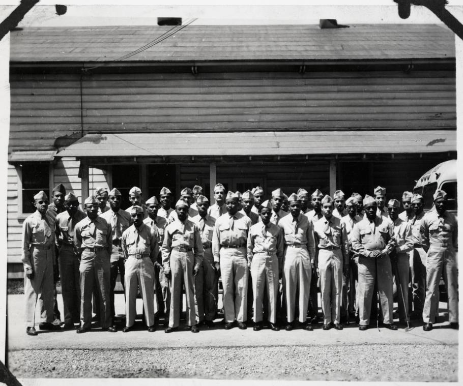 Black and white photo of approximately 30 Black men in uniform, posing in front of a wood building with an awning.
