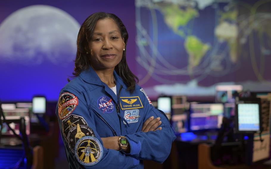 Stephanie Wilson stands with her arms folded, wearing a blue jacket with many space mission patches.