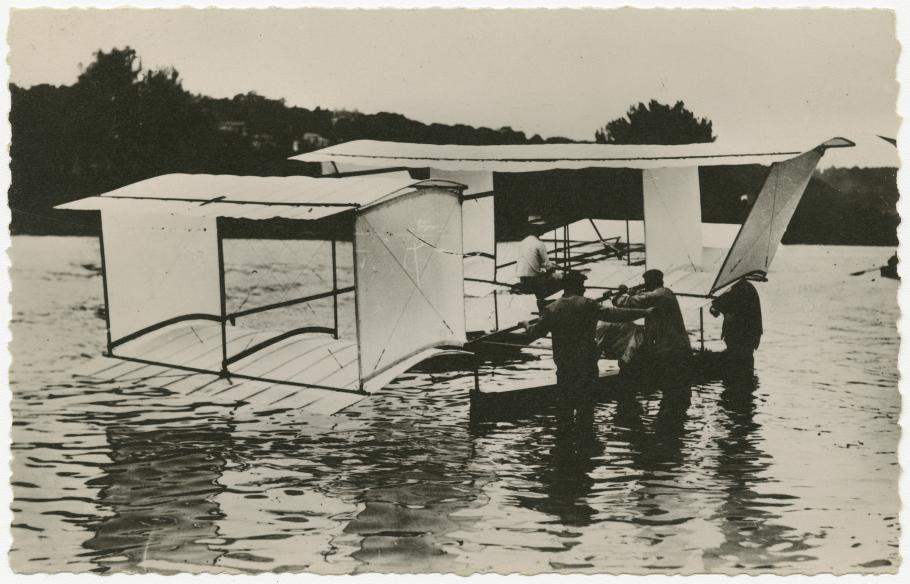 One-half right rear view of the a Blériot floatplane on the water. Men stand in the water next to it.