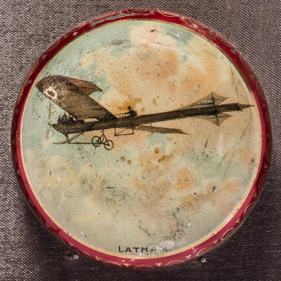 Circular metal box with monoplane and pilot in a cloudy sky painted on the lid. Image surrounded by narrow gold border. Sides and bottom metallic red.