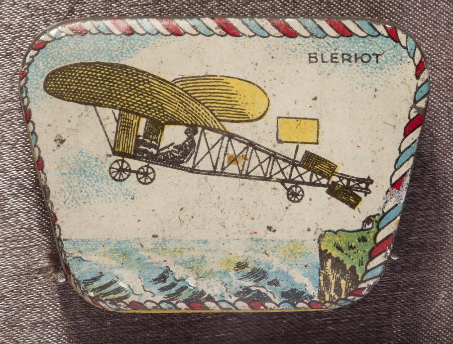 Small metal trapezoidal box depicting Bleriot monoplane ascending into the sky, flying to the viewer's left. The edge of a grassy cliff is visible on the right, with waves below the plane. "Bleriot" in black block letters upper right. Bordered in red, white, and blue rope.
