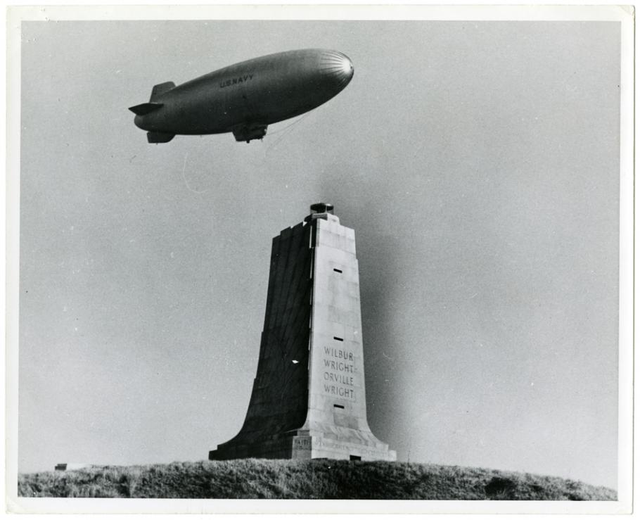 One-half right front view of a U.S. Navy airship in flight over the square column-shaped Wright brothers memorial, which has both brothers names inscribed on it.