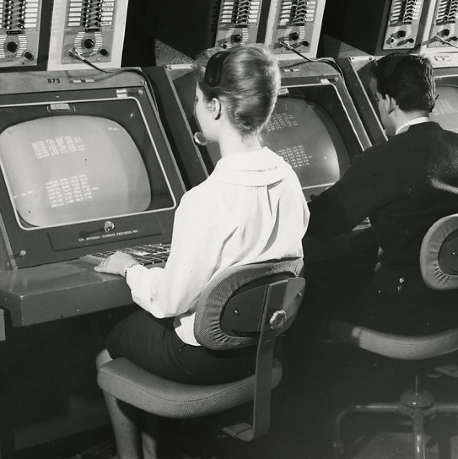 Two people sit at computer terminals, facing the screen working. 