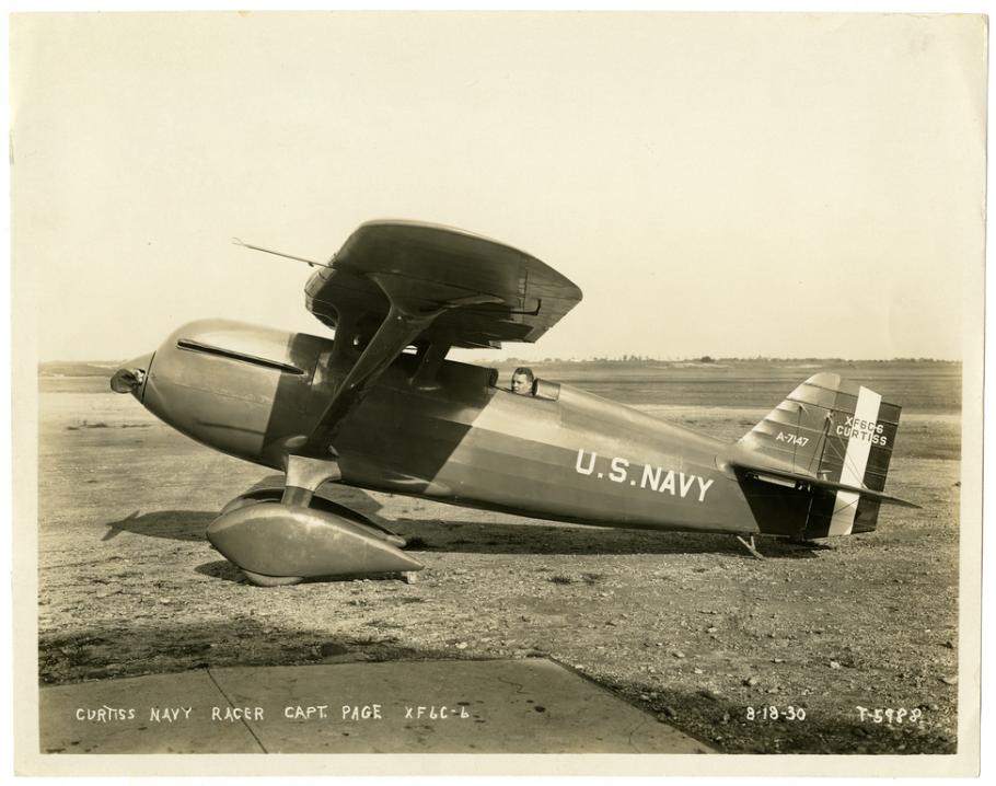 Sepia-toned photo of the left side view of a person in a Curtiss racer monoplane on the ground. The aircraft has U.S. Navy written on the side. 