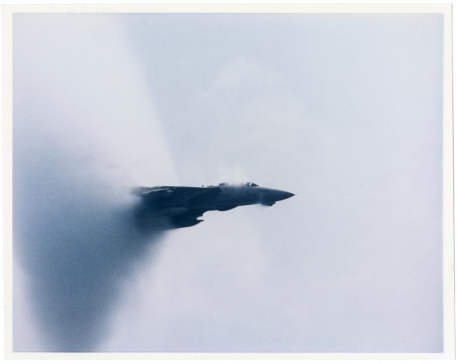 A jet aircraft emerging from a cone shaped cloud of vapor in the air. 