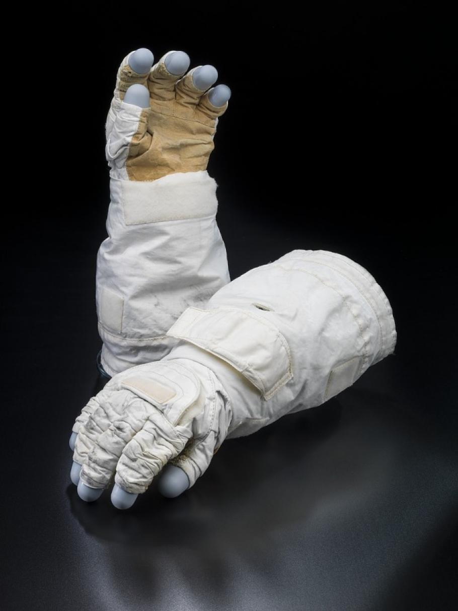 Two gloves to a space suit. One lies down and one stands up. The gloves are made of a white material, with velcro sections, the finger tips appear to be blue plastic. 