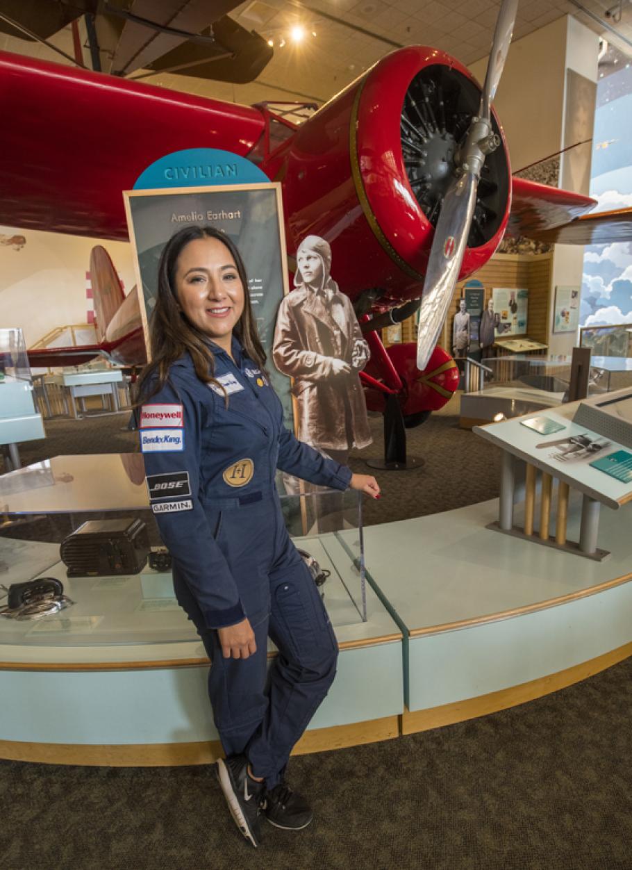A woman in a flight uniform poses next to a bright colored early aircraft in a museum setting.