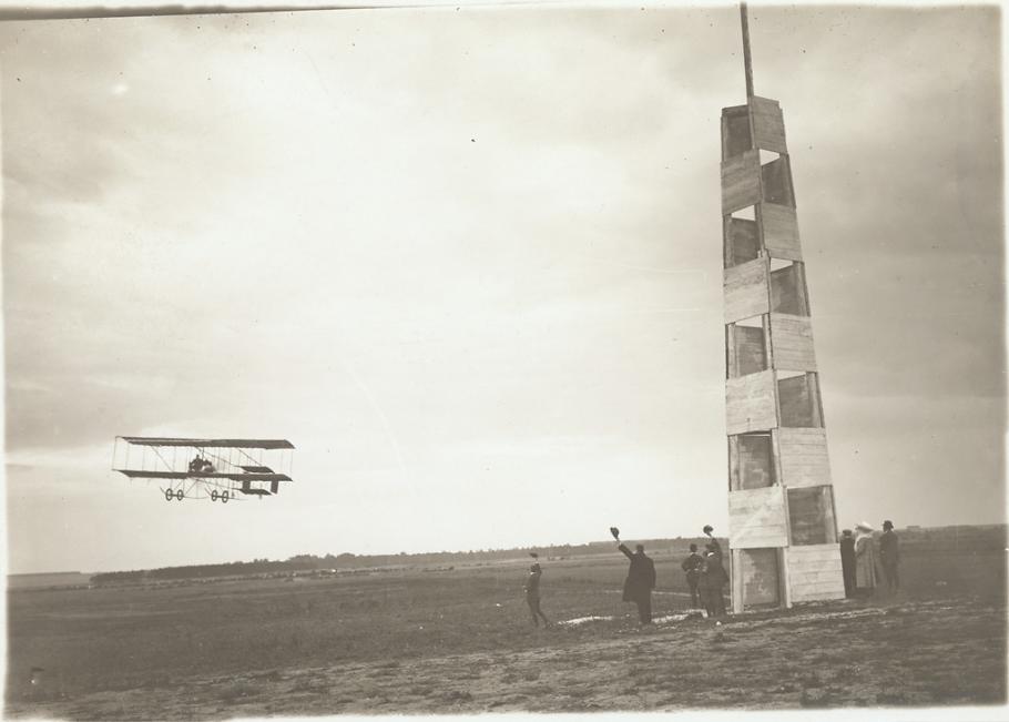 Farman-type biplane rounds a pylon in a field as a handful of people look up, some waving their hats in the air.