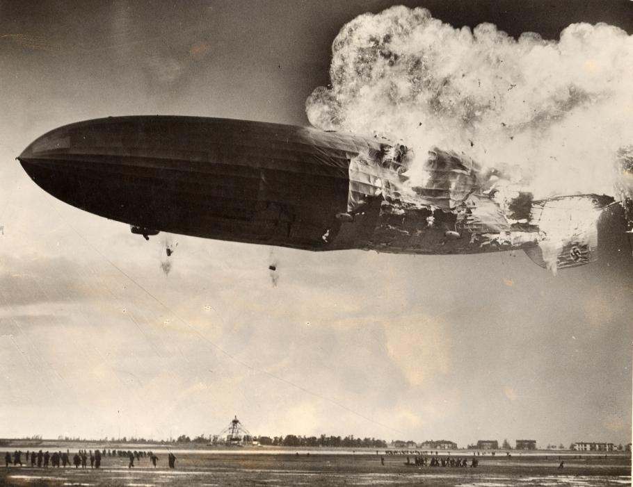 An airship is seen with the back half of its body on fire as it is still in the air.