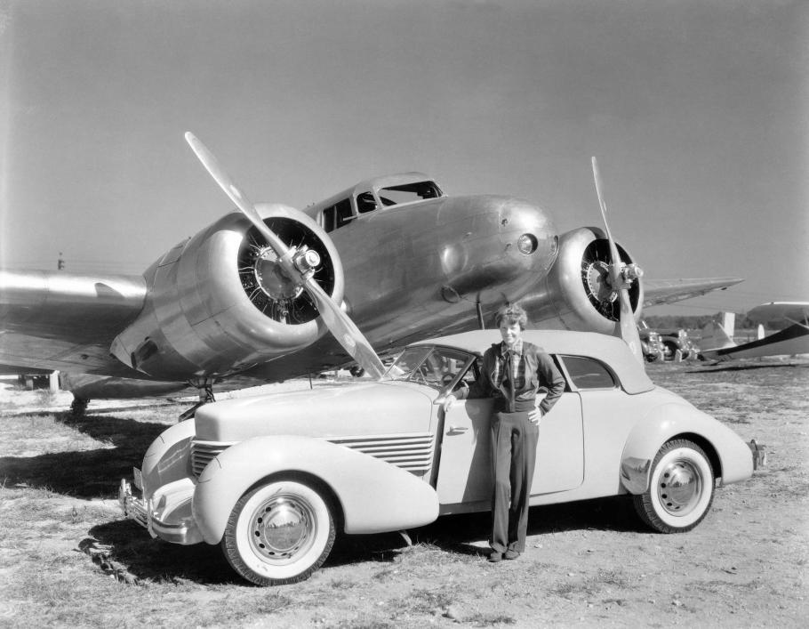 A wideshot of a woman standing next to a car with a aircraft in the background.