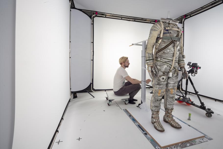 A man sits in studio where a spacesuit stands in the foreground.