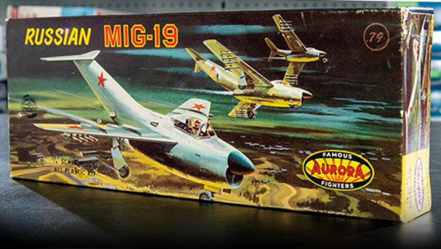 A plastic model airplane kit sits on a table, with artwork on the box depicting a Russian MiG-19.