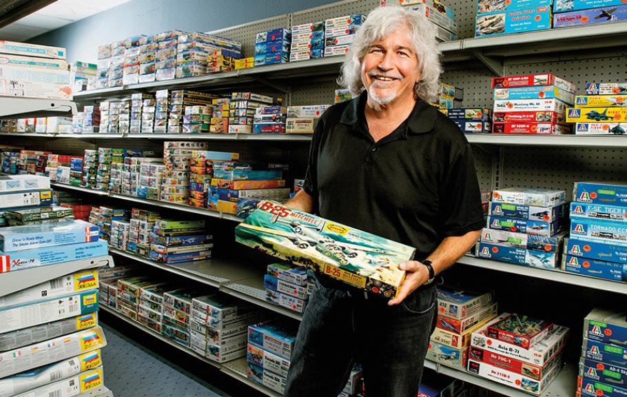Jeff Garrity,—a 65-year-old man sporting long hair and a goatee, smiles while surrounded by shelves filled with vintage plastic airplane models.