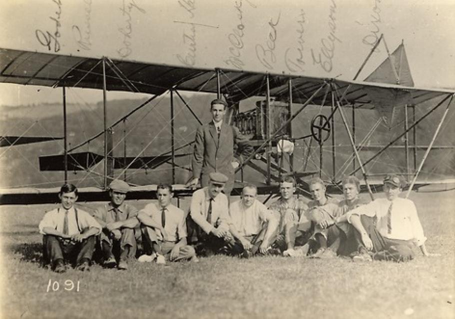 Nine people sit in a row on the ground, another person stands behind behind them in the center. They pose in front of a biplane.