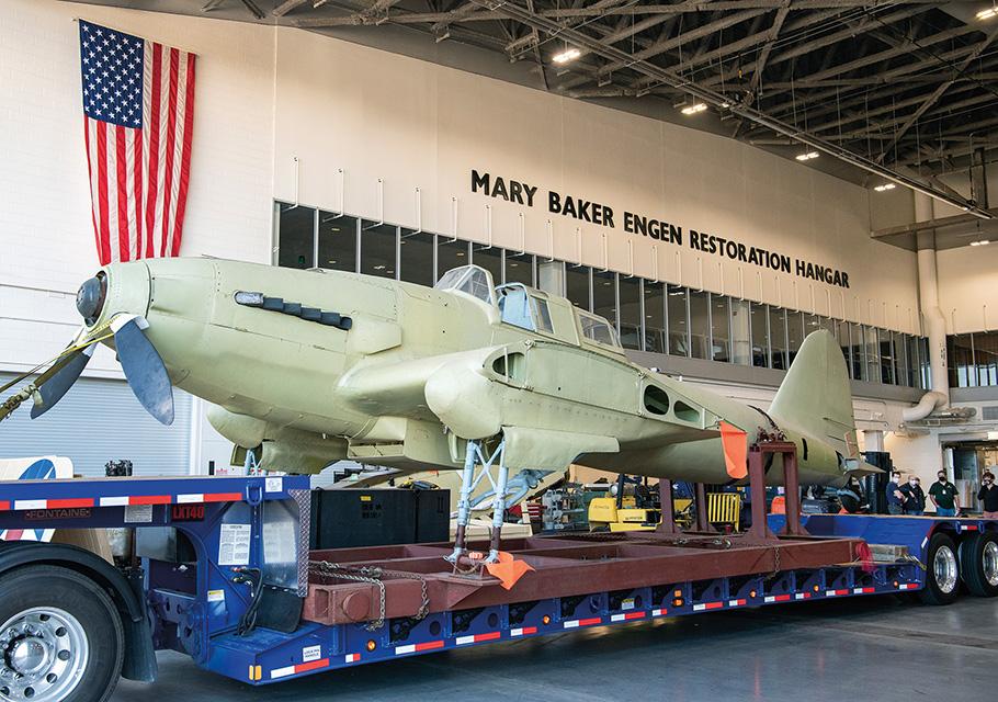 An IL-2, with its wings removed, is mounted on a flatbed truck, beneath a large sign that reads: Mary Baker Engen Restoration Hangar.