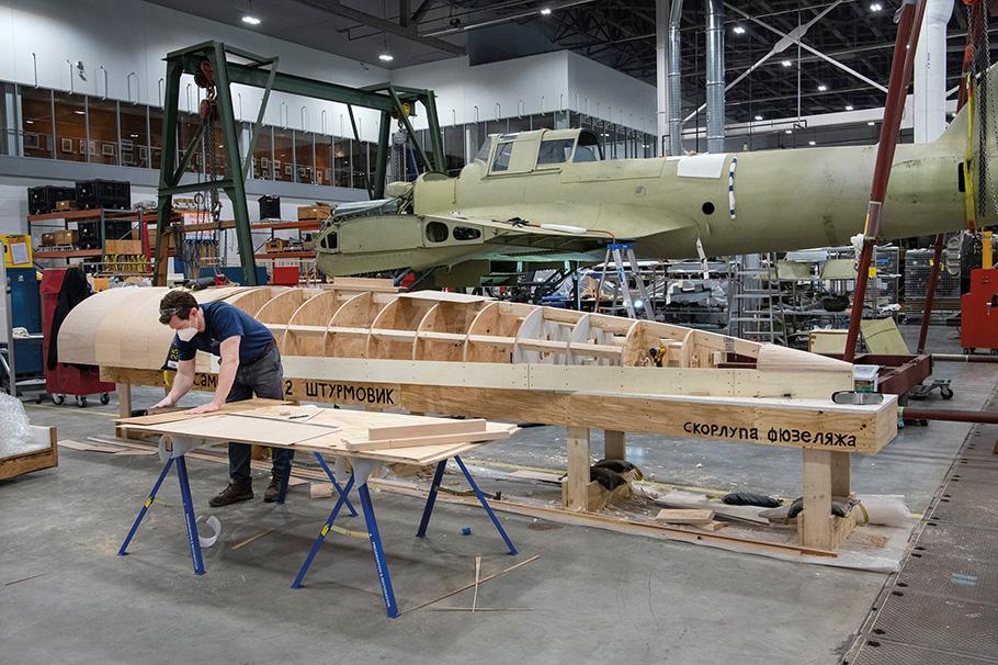 Museum Specialist Jay Flanagan is hunched over a piece of wood on a work bench; behind him sits the skeleton of a wingless IL-2 aircraft wooden fuselage that he is building.