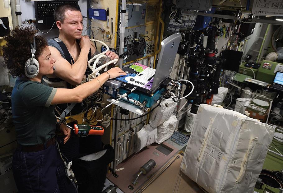 Two astronauts are conducting medical experiments aboard the space station, standing amid cluttered equipment. A woman wearing a t-shirt looks on as a man holds onto a tube that is attached to a laptop.