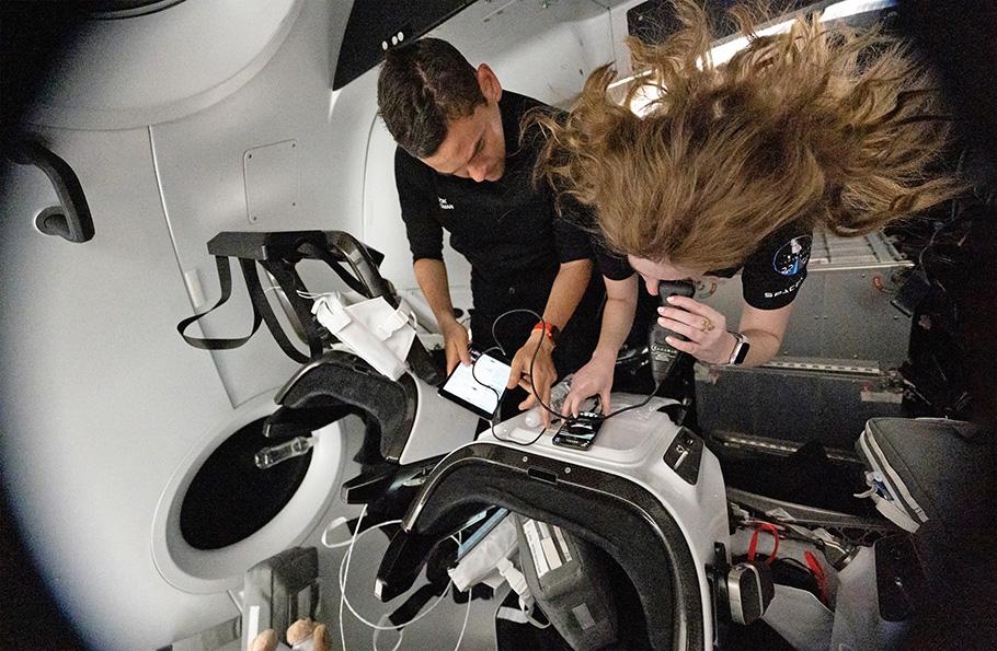 Two civilian astronauts conduct medical research in orbit as part of the Inspiration4 mission. A young woman, her hair floating above her, holds a small medical instrument against her eye. The instrument is attached to an iPhone.