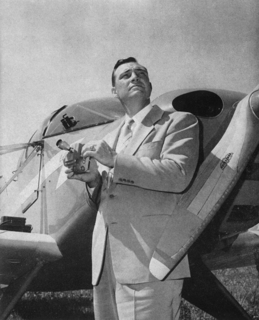 Black and white image of a man standing in front of an airplane, holding an object in his hand, and looking to the right.