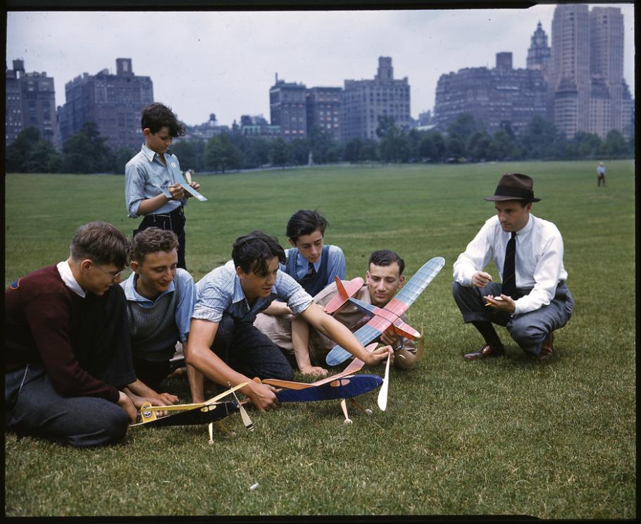 A group of six young men of various ages gather around three model airplanes in a park with a city skyline in the distance. To the right is an adult crouching on the ground next to them with what seems to be either a stopwatch or notebook. 