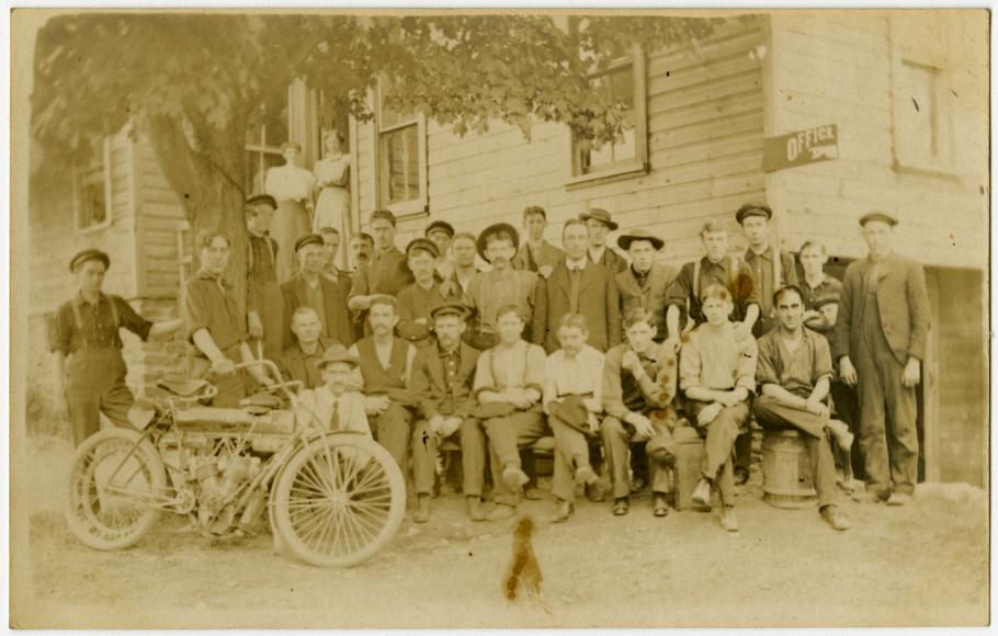 A large group of people pose in front of a building in this sepia-toned photo. A motorcycle is propped in front of the left side of the group.