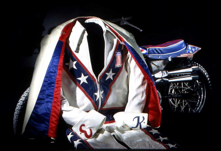 A red, white, and blue colored jumpsuit and cape propped in a sitting position. In the background is a partial view of a red, white, and blue motorcycle.