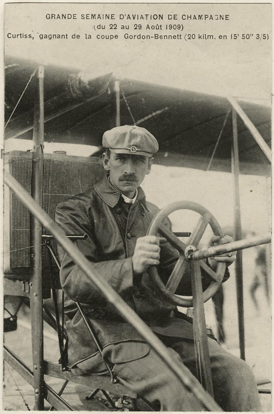 A man sits behind the steering wheel of a biplane, looking toward the camera. The photo looks old and is dated August 1909.