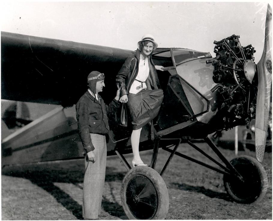 A woman looks toward the camera as she climbs through the door of an aircraft. A man to the left, Clyde Cessna, assists her.
