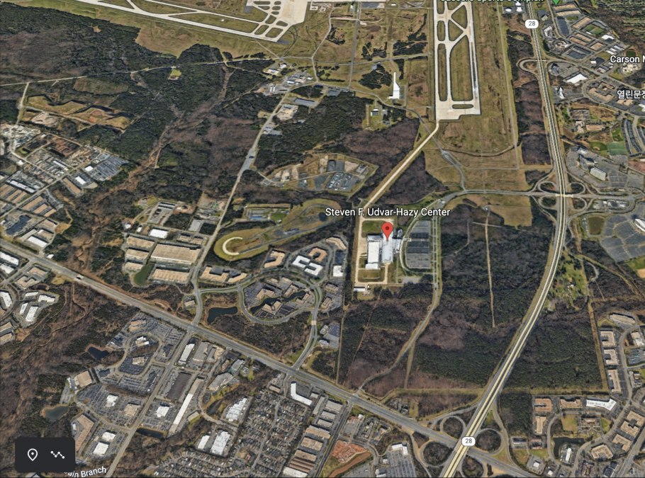 An overhead view of an aircraft hangar surrounded by a parking lot, roads, and many many trees. 