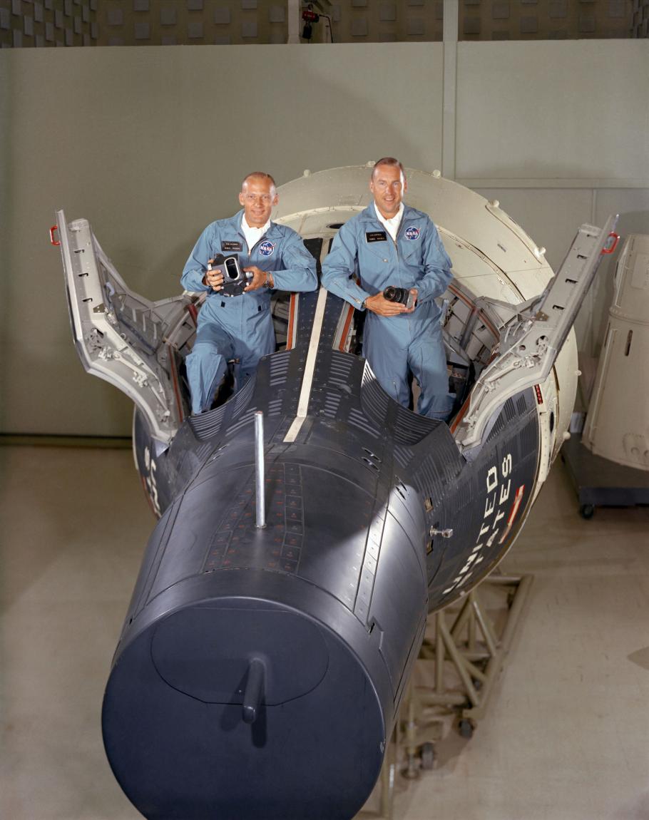 Two men in flight suits pose standing in open hatches of a Gemini spacesuit.