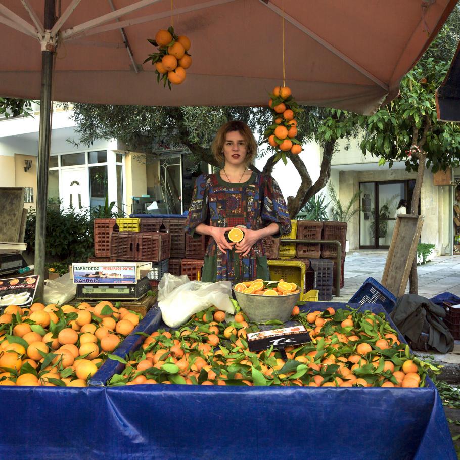 A woman stands behind an outdoor fruit stand, holding a piece of fruit.