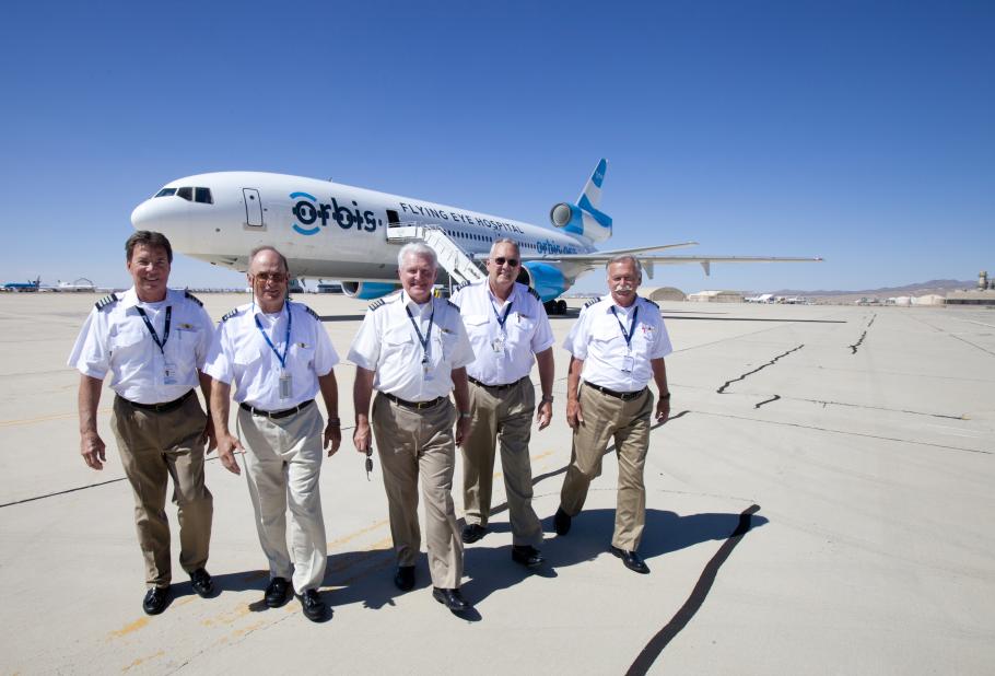 Five pilots walking toward the camera shoulder to shoulder. The Orbis Flying Eye Hospital can be see in the background.
